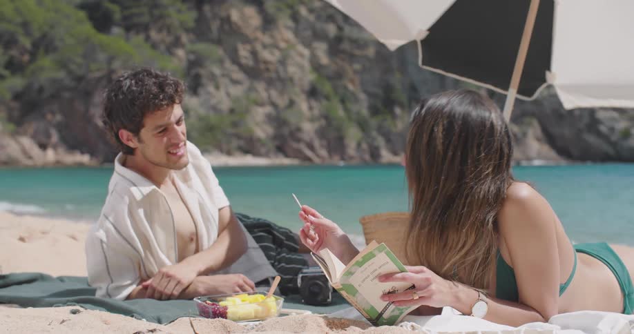 A young couple seated on a beach towel by the shore eating fruit