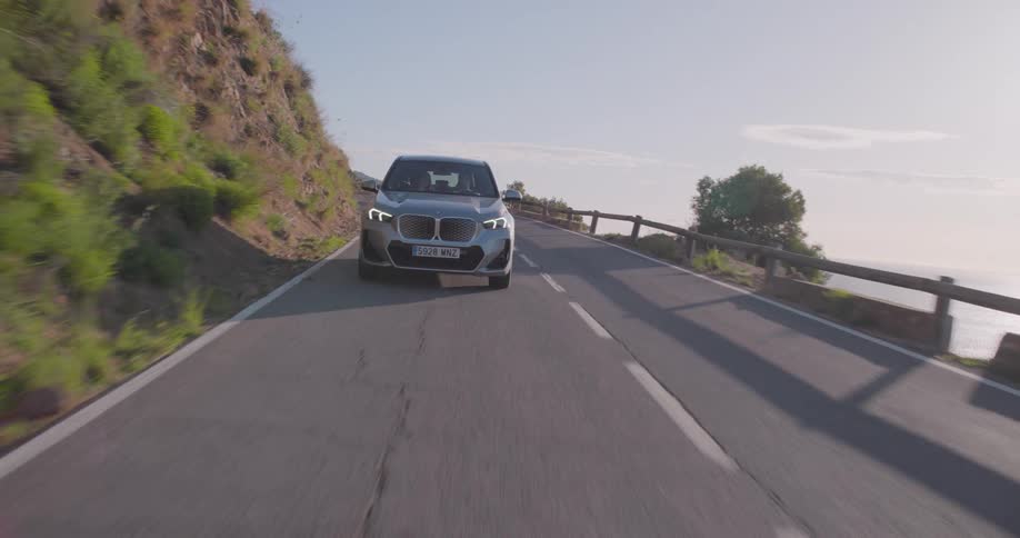 A man and woman travel in a car down a coastal highway on a mostly sunny day