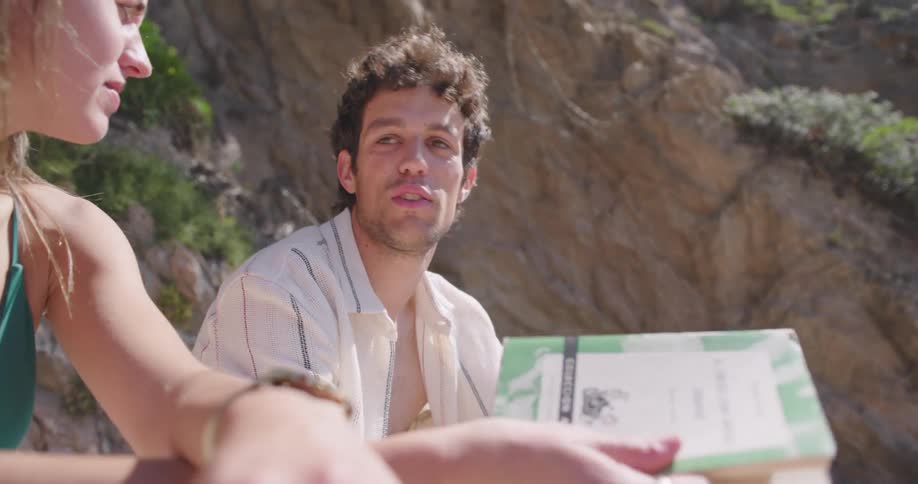 Couple sitting on a beach with a woman showing a book and running her hand through her hair