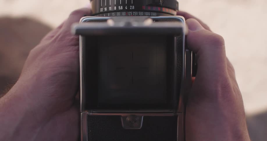 Hands holding a vintage camera with a woman on a beach seen in the viewfinder