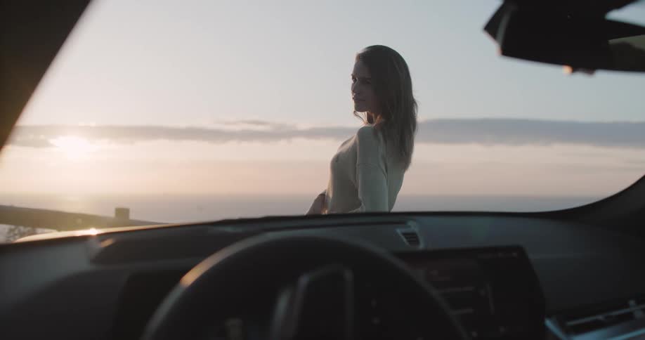 Woman reclined on a car with a sunset or sunrise visible through the windshield over the ocean