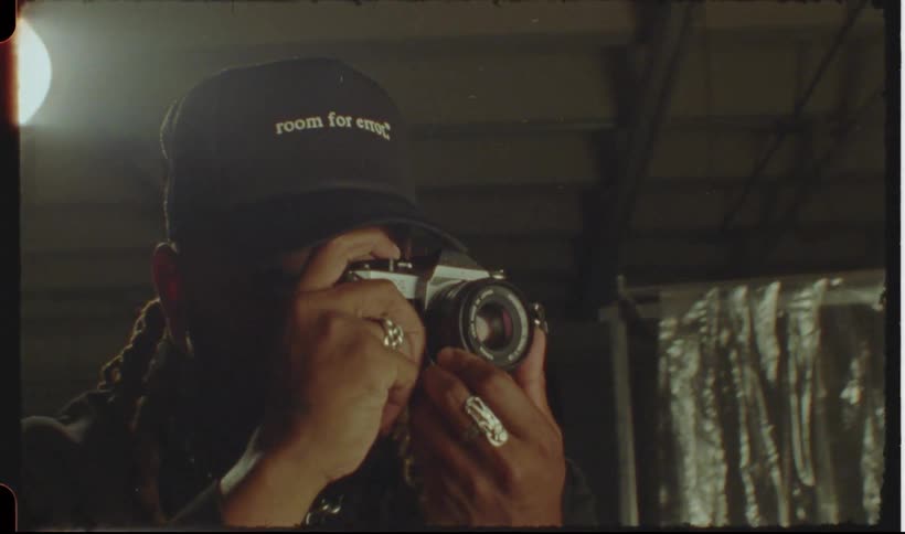 A manwith braided hair wearing a cap is taking a photograph using a vintage camera