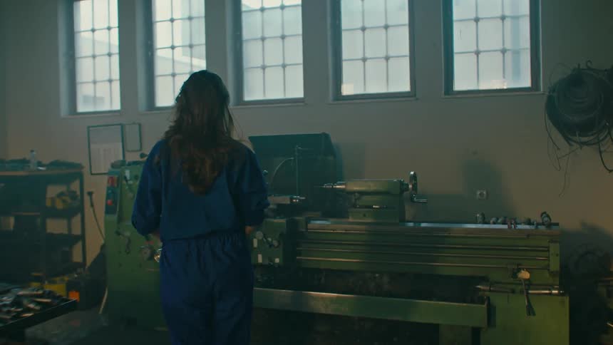 A woman in blue coverall working on a large industrial lathe in a workshop