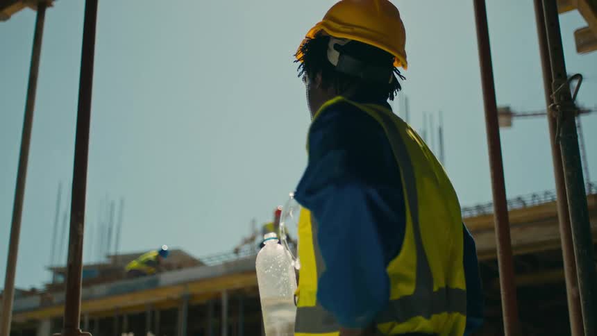 A construction worker in a yellow hard hat and reflective vest is drinking from a water bottle