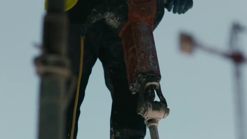 A construction worker in a yellow reflective vest and orange hard hat is operating a jack hammer on a construction site