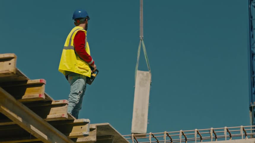 A construction worker in a yellow vest and blue helmet on wooden scaffolding