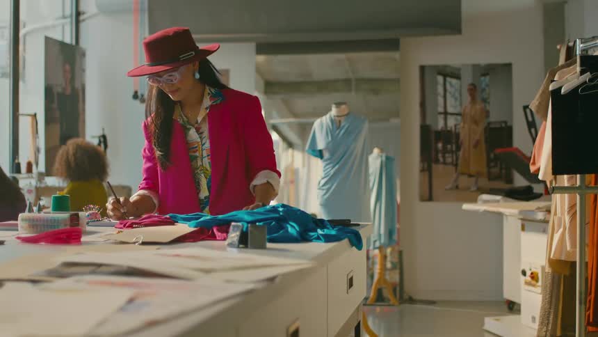A fashion designer in a red hat and pink jacket working on blue fabric at a sewing station with tailoring tools