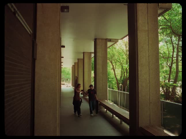 Two young friends walk inside a building surrounded by trees carrying skateboards