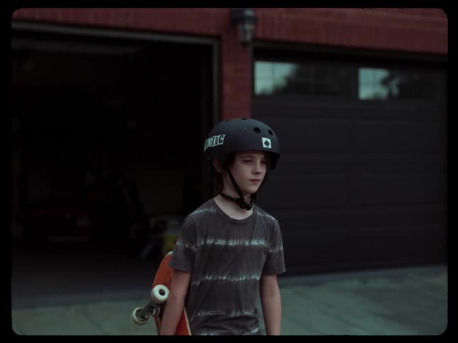 A boy with a skateboard stands in front of a garage door wearing a helmet and a dark tshirt