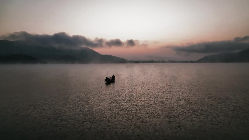 Aerial panorama of two people in a canoe on the calm lake with the silhouette of mountains around in a reddish sky
