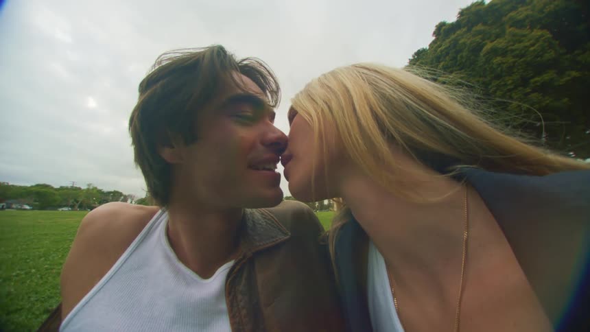 A man in a white undershirt and brown leather jacket leans in to kiss a blonde woman in a white top and black jacket who is sitting on a green lawn