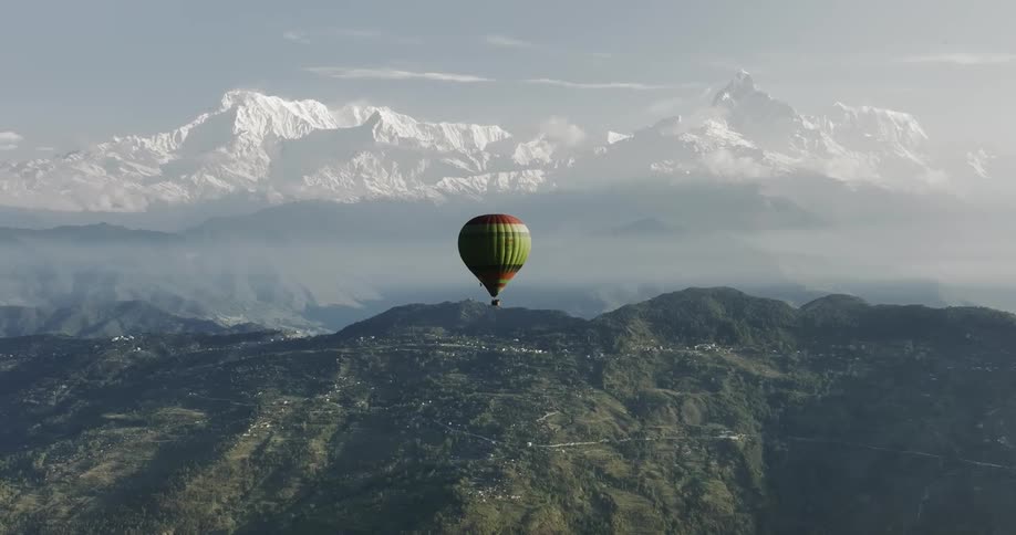 A hot air floating in front of snowcapped mountains and over green hills
