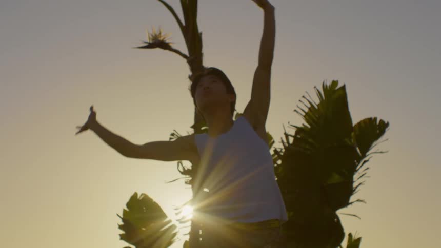 Man in a white tank top dancing and throwing his arms up and to the sides while standing in front of a bush and sunlight reflecting
