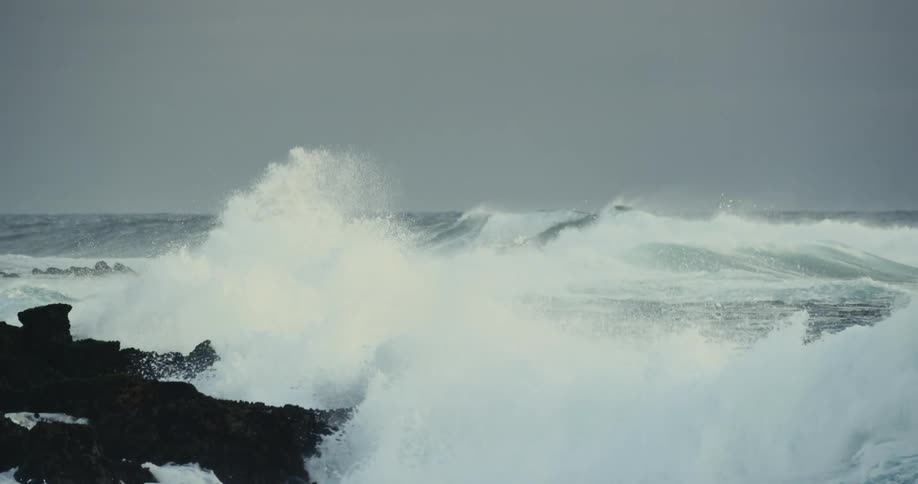 Big waves roll onto black coastal rocks under a gray overcast sky