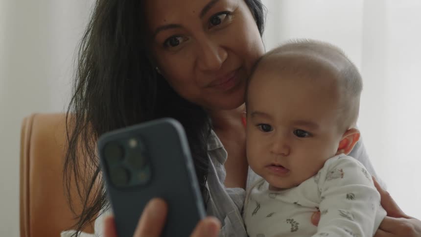 A mother holding her baby while looking at her smartphone making a video call and kissing the baby