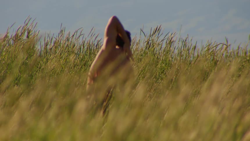 A young shirtless man dances on a field of tall grass on a sunny day