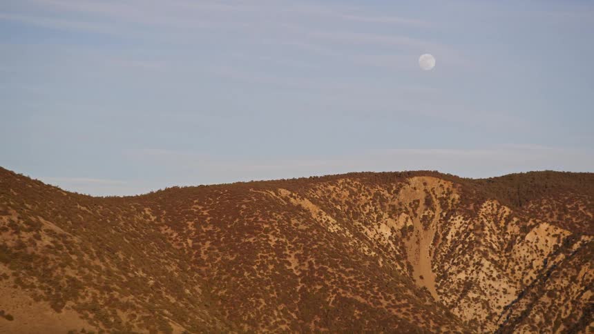 Aerial panorama of mountain ranges with vegetation against a blue sky during the day and with the moon visible