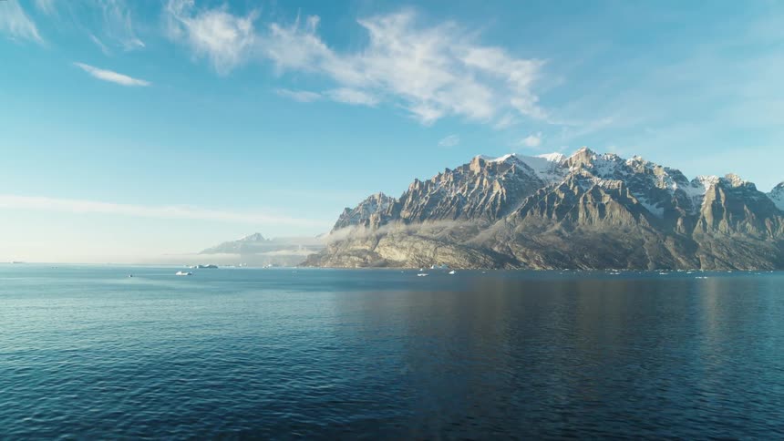 A panoramic view of a rugged mountain range beside a calm sea with small icebergs and clear blue skies