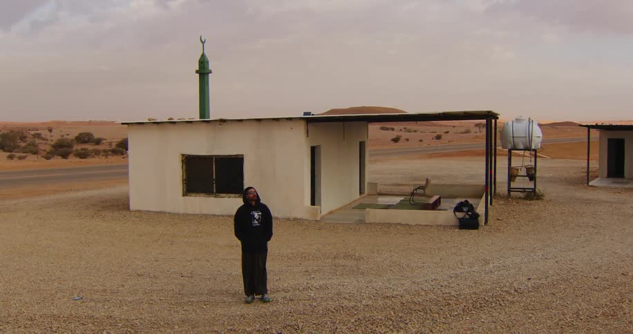 Man with sweatshirt and hood walking in a desert environment next to a small house