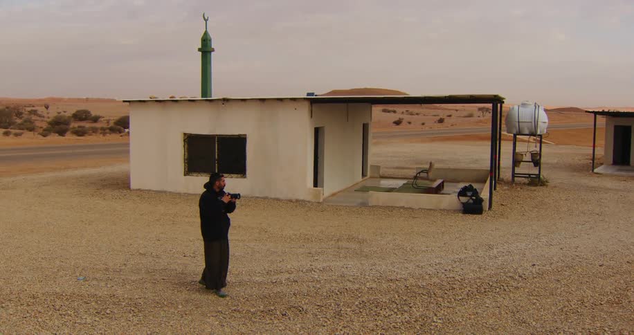A man in black clothes is using a camcorder to film the environment and then films a small house behind him