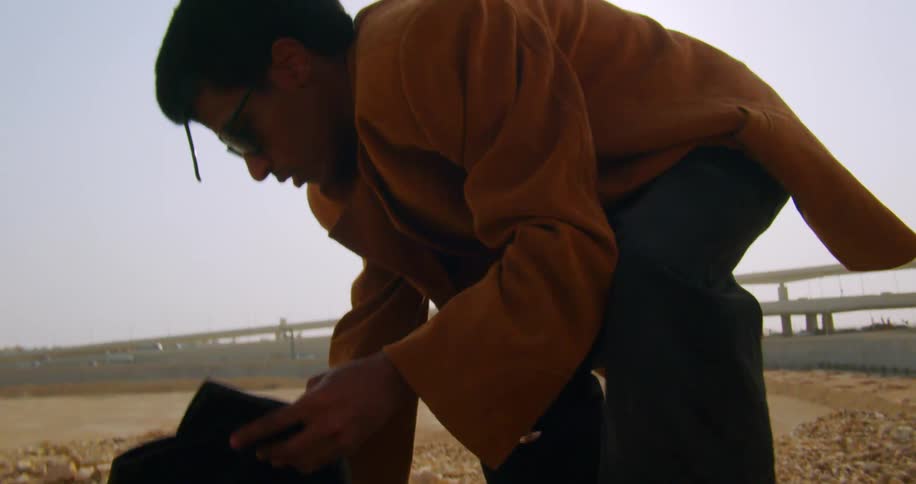 Man wearing a coat and cleaning leather boots while outdoors next to an overpass