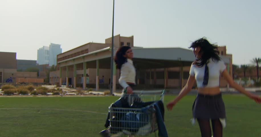 Two laughing girls run while rolling a grocery cart on a field