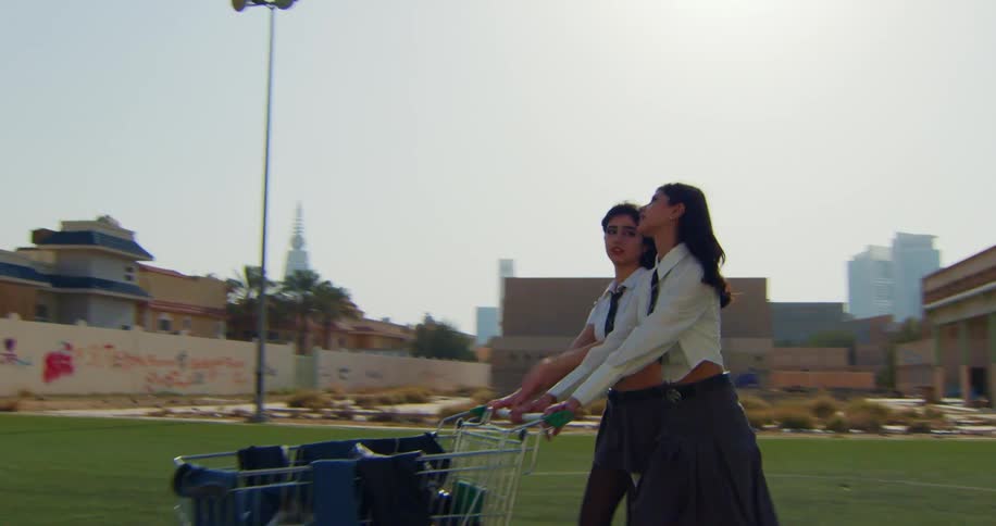 Two young models in student clothes walking with a shopping cart on artificial grass in an outdoor location