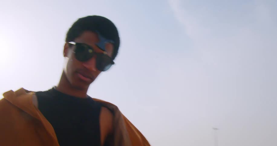 A man in sunglasses and an orange jacket walking near a highway and he is looking straight ahead and fiddling with his hair and glasses