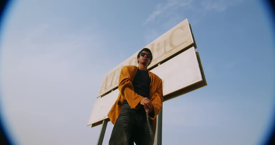 Man with sunglasses and a long suede coat biting a chain in an openair location in front of a commercial sign