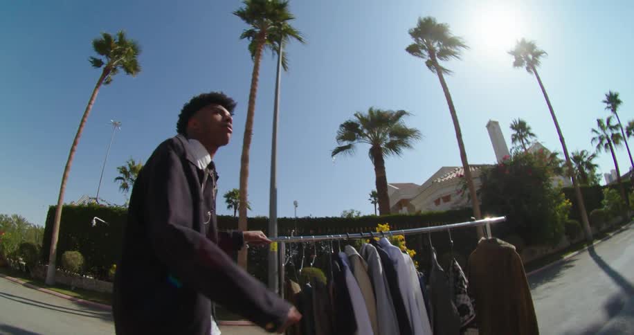 A young man pushing a clothes rack down a street with palm trees on both sides of the road