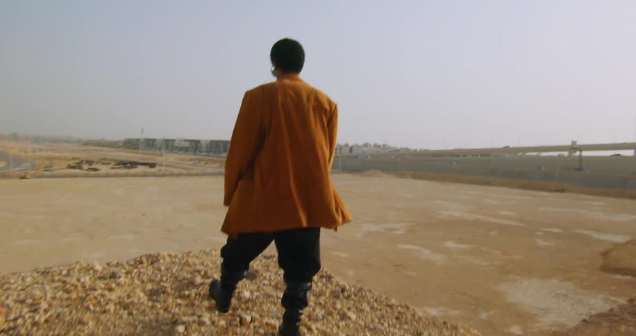 A person in a brown jacket and dark pants stands alone in an empty barren construction site facing away from the camera with cranes and and a bridge visible in the background