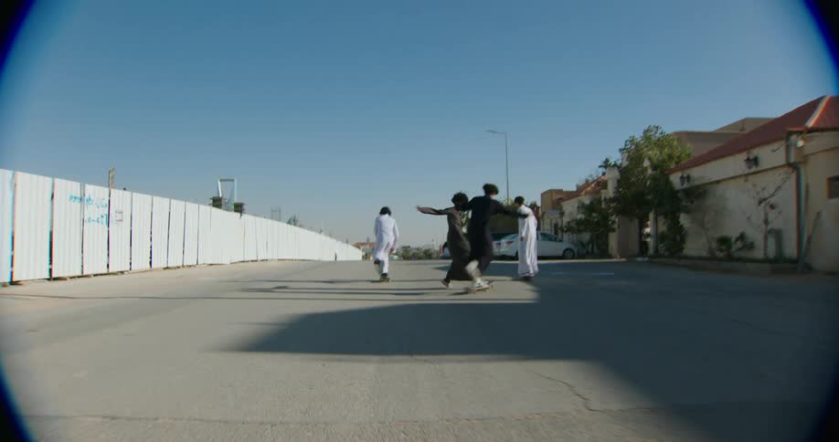 A group of friends wearing long robes skateboarding on a street with a high fence on one side under a clear blue sky