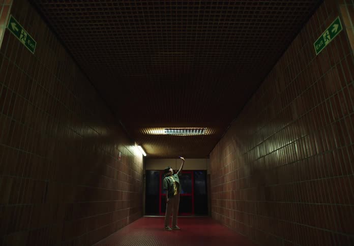 A person stands in a dimly lit hallway with tiled walls facing up with an arm outstretched