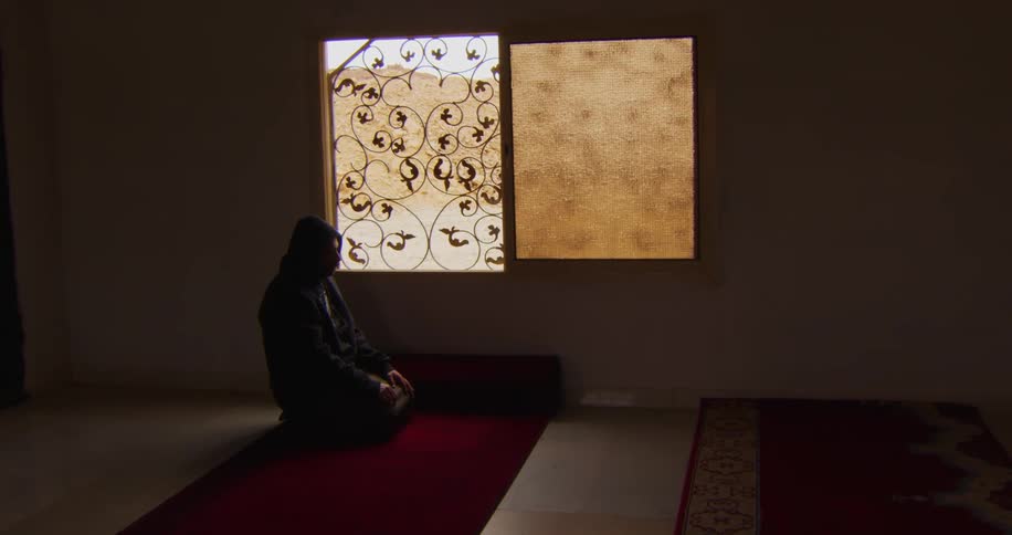 An individual sitting on a red carpeted floor leaning against a wall next to a window with ornate iron grills