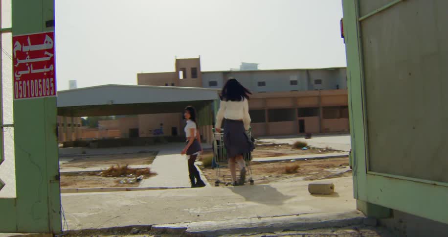 Two teenagers wearing schoolgirl outfits entering an abandoned lot through an open gate