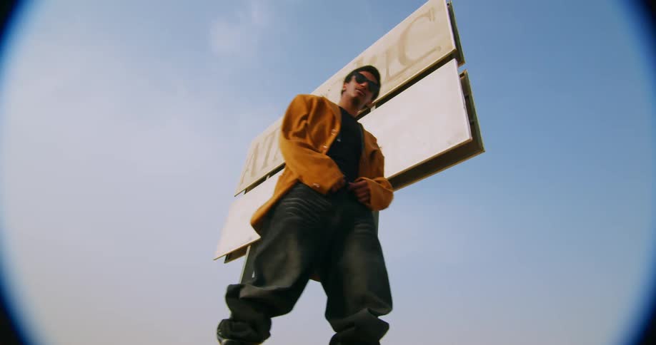A young man in an orange jacket gets up from the ground where he was sitting and strikes poses in front of a billboard
