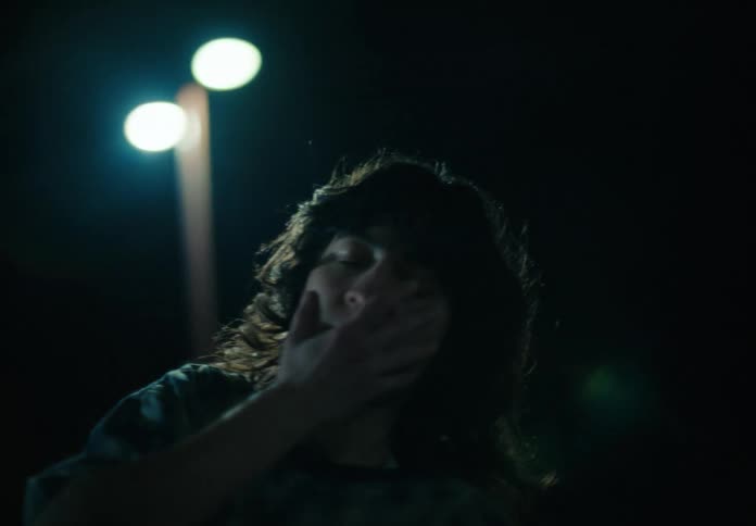 A woman with wavy hair and a blue tshirt dances against a streetlight on the street at night