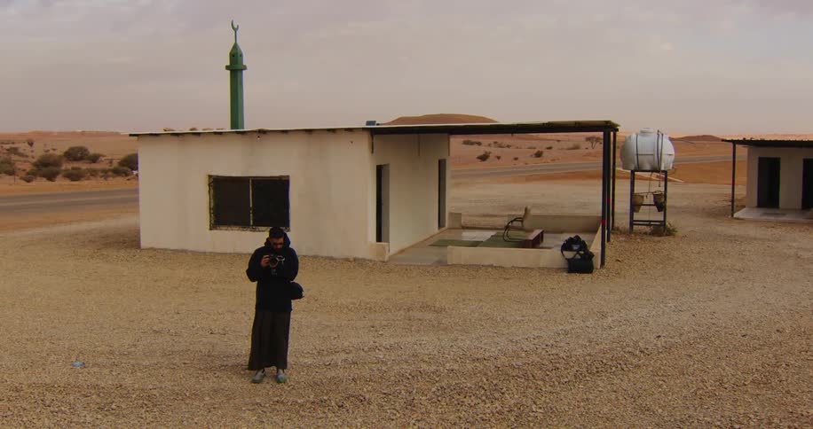 A person standing in a barren area with a small building featuring a green minaret taking pictures