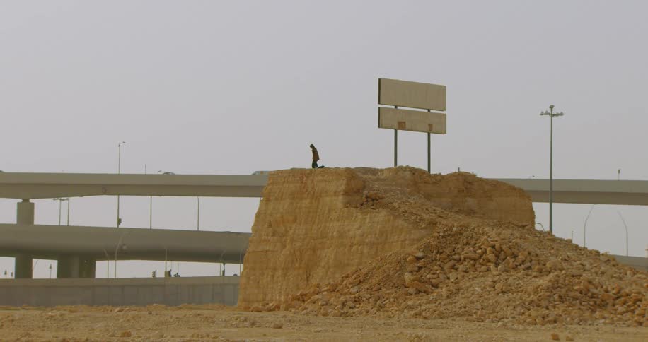 A distant man stands on a mound of dirt next to a billboard with cars crossing overpasses in the background