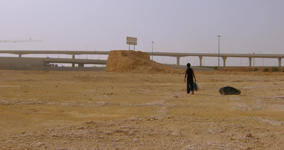 A man wearing a black undershirt and pants who is carrying a suitcase away in a construction area