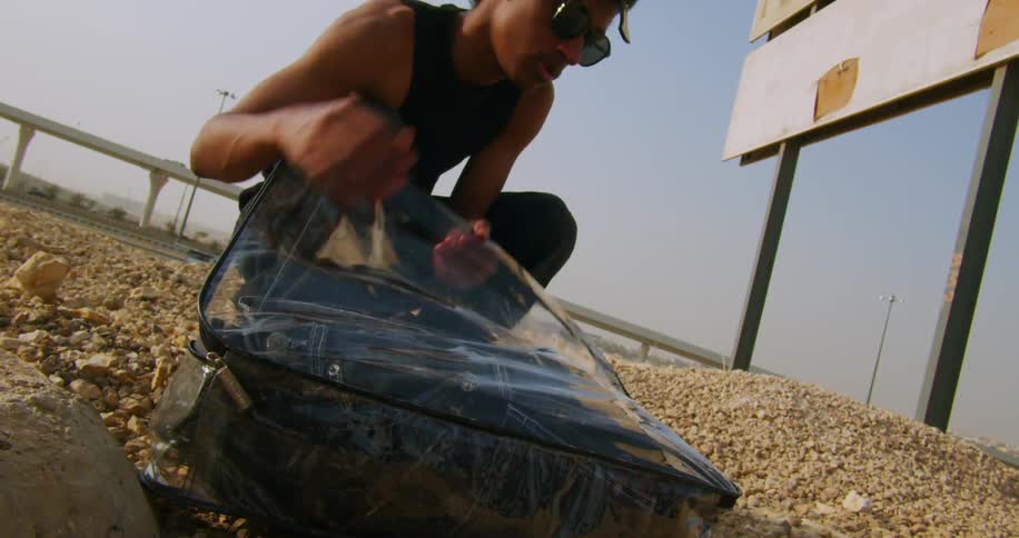 A man wearing sunglasses a black undershirt and pants walks up to a suitcase lying on the ground and opens it in the construction area