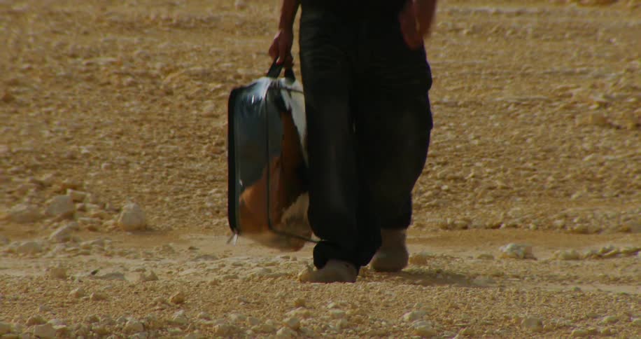 A man walking on a ground of gravel carrying a bag of clothes shaped like a briefcase