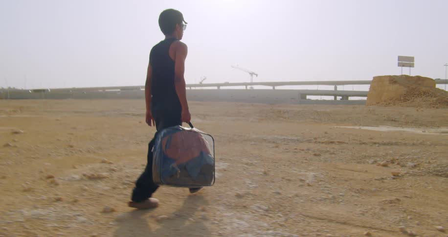 Young man carrying a clear suitcase with clothes walking across a field towards the highway