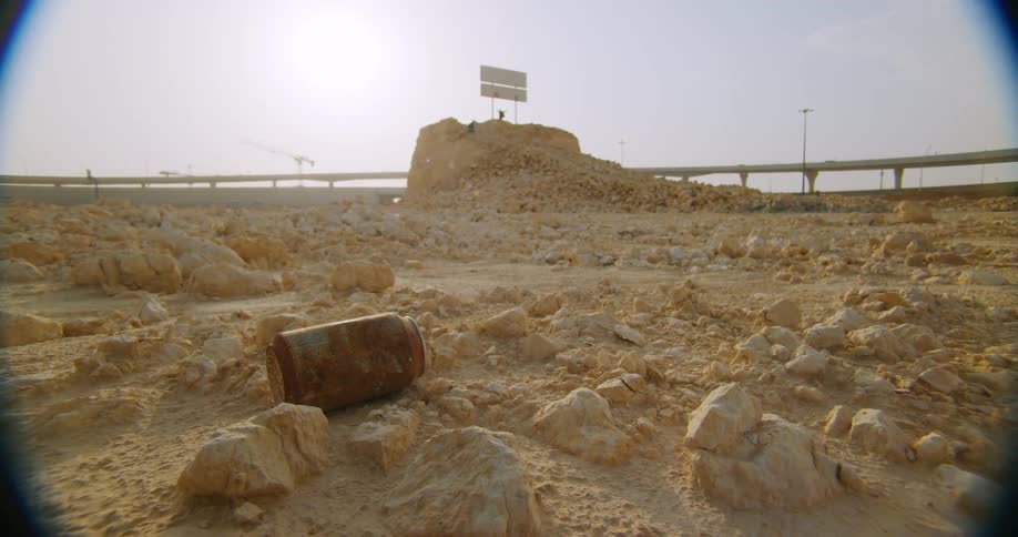 A soil of sand and stones with a rusty metal can and in the background there are cars passing on a road