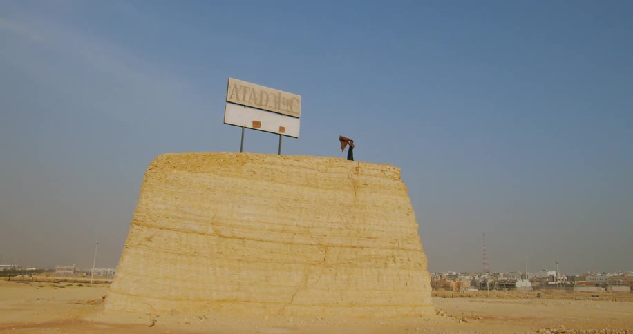 Person on top of a hill next to a sign in a desert terrain with buildings in the background