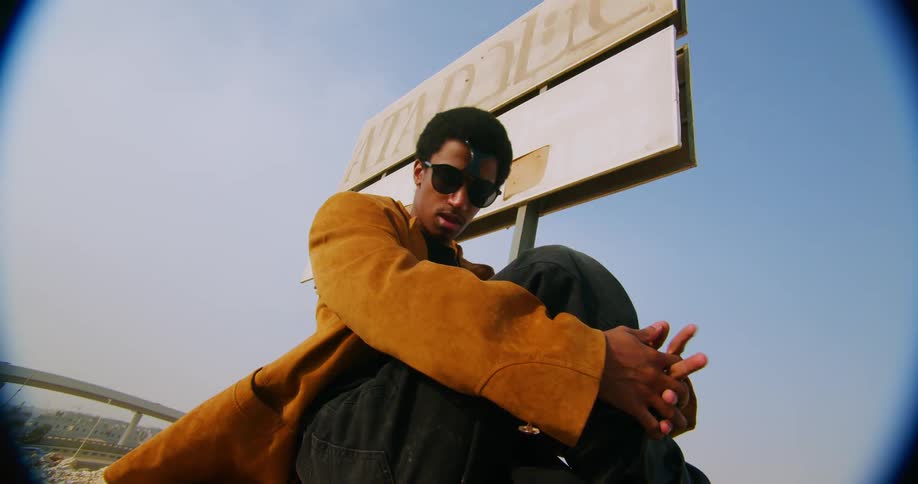 A man in sunglasses wearing an orange jacket is posing looking forward and fidgeting with his clothes while crouching down