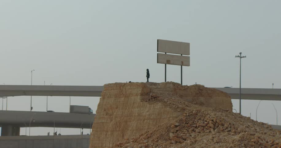 A person stands on a large dirt mound situated near overpasses