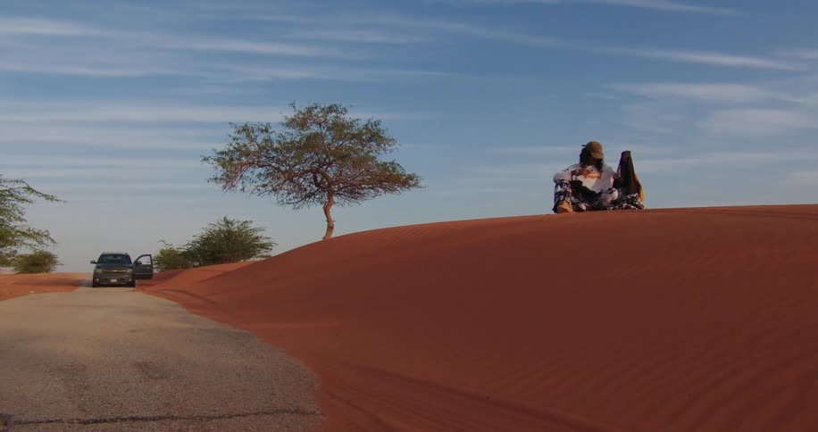 A disheveled man sits on a sand dune sorting clothing