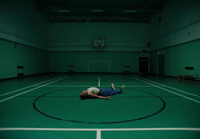A person is lying on their back in the center of an indoor basketball court with dim lighting