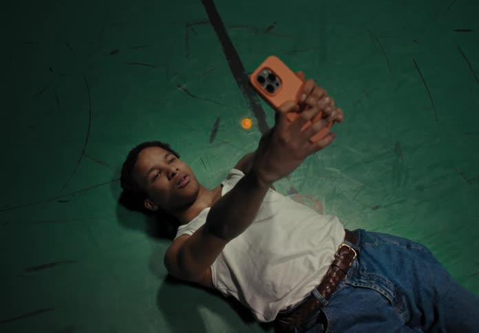 Man wearing a white undershirt jeans and a brown belt lies on the ground taking a selfie with a smartphone